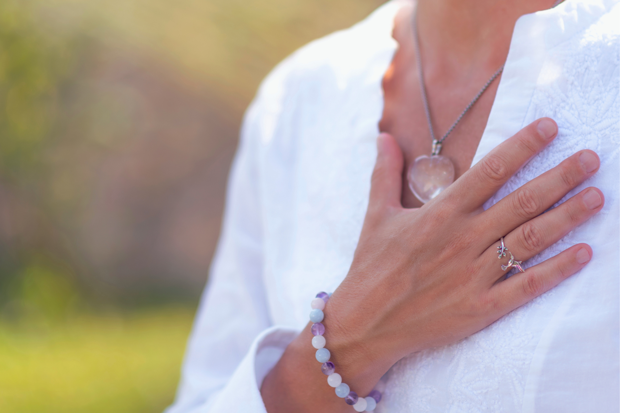 Frau in weißer Bluse mit blau-lila-weißem Perlenarmband und Kristallherzkette hat ihre rechte Hand auf ihrer Herzgegend liegen